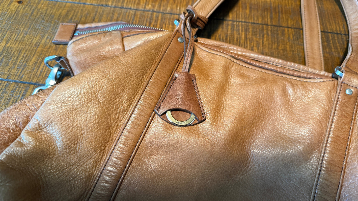 A close-up of a tan leather shoulder bag with a pebbled texture, featuring a zippered pocket with a leather pull tab and a brass-tone oval ring detail, resting on a dark wood floor.