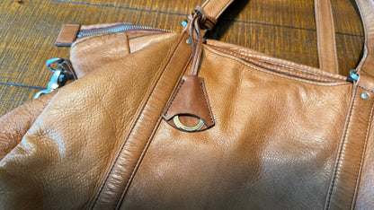 A close-up of a tan leather shoulder bag with a pebbled texture, featuring a zippered pocket with a leather pull tab and a brass-tone oval ring detail, resting on a dark wood floor.